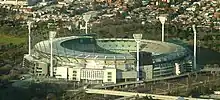 Aerial view of outdoor sport stadium. Erected around the stadium are six tall flashlights. The stadium are surrounded by houses, parklands and railway lines.