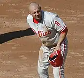 A man in a gray baseball jersey having just removed his red batting helmet at first base. His jersey reads "Phillies" in white and red script and has a number "8" on the left sleeve.