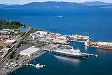 One large ship and one small ship are docked at a port. There is a large expanse of water with another small ship approaching and mountains in the background.