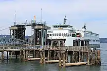 A ferry boat stopped at a wooden pier with a bridge, pilings, and a tower.