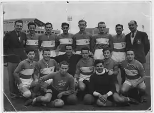 A black-and-white photograph of an association football team. A row of eight men stands at the rear, six in dark-coloured soccer jerseys with a light stripe horizontal across the centre, and one on each end in dark-coloured suits. In front of them sit eight more players. In the background a tall floodlight and two grandstands can be seen.