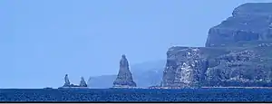 A large sea stack and two smaller ones stand off a coast of  high cliffs. There is a deep blue sea in the foreground and two further peninsulas with high cliffs beyond.