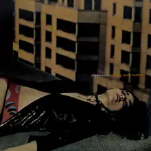 Madonna laying down at the corner of a rooftop, with yellow buildings present behind her. She wears a black shining jacket.