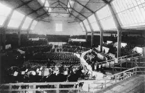 Darkened interior of a large hall with two rows of high windows along each side. It is possible to discern a seated orchestra in the foreground, with mass choirs in the background.