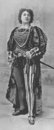 A white woman with dark curly hair, standing, wearing a theatrical costume with voluminous sleeves and striped tights