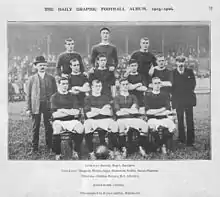 A black-and-white photograph of a football team lining up before a match. Four players, wearing dark shirts, light shorts and dark socks, are seated. Four more players are standing immediately behind them, and three more are standing on a higher level on the back row. Two men in suits are standing on either side of the players.