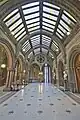 The landing outside the Great Hall, Manchester Town Hall, showing the mosaic floor, and skylights providing light not just for the landing but to the adjacent corridor, the column shafts are of grey or red granite, the arches are of stone, the dado has a pattern formed of plain ceramic tiles, the upper walls are clad in buff coloured terracotta, interspersed with thin bands of blue terracotta, the doors on the right lead into the Great Hall