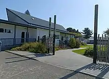 a wooden single level building in the sunshine, with a gateway in the foreground and grass and plants