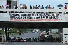 People stand on a highway with a banner hanging down that reads: Menhuma mulher deve ser presa, maltratada. humilhada ou morrer por fazer aborto.  Prente nacional pelo fim da criminalizacao das mulheres e pela legalizacao do aborto.