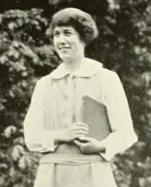 A young white woman, standing outdoors, smiling, wearing a light-colored dress and holding a book under one arm; her hair is cropped to earlobe length, with bangs