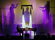 A blonde woman sits atop a piano and sings. A long, white cloth hangs around her while a silhouette behind the woman shows two male figures as if holding the white cloth.