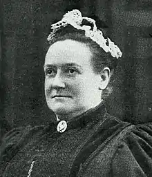 A white woman wearing a lace cap atop her hair, and dark dress with a cameo pin at the collar.