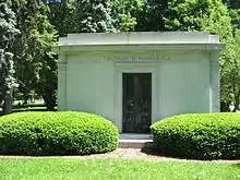 A large stone tomb with a metal gate in the center of the tomb, and with carved columns at the corners meeting a crest which circles the top of the building. Two large evergreen bushes obscure the lower portion of the tomb which sits in a grassy meadow.