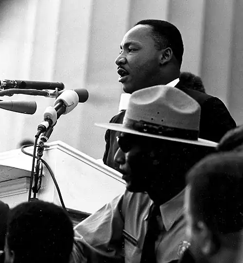 In a black-and-white photograph, a middle-aged African-American man stands in front of multiple microphones. In the foreground is a middle-aged white man wearing a US police uniform.