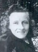 The smiling face of a white woman, wearing a dark garment fastened high on her neck, and her hair dressed back from her forehead.