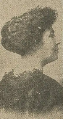 A white woman photographed in profile; her dark hair is in an updo; she is wearing dark garment