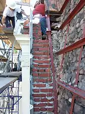 Cross-section of brick masonry exterior wall with cast iron cladding. Workers set new brick courses against the existing masonry back-up.