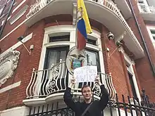 Garrett in 2016, standing outside the Ecuadorian Embassy in London, where WikiLeaks founder Julian Assange had received asylum, holding a sign saying "You're not even a Wiki"