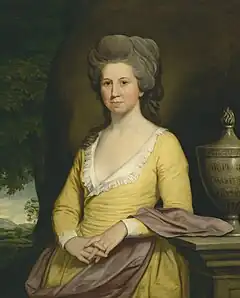 Painting of woman, at three-quarter length, wearing a yellow dress with a plunging neckline, which has a white lace trim. Her hands held together in front of her, with fingers entwined. She leans on a stone balustrade, upon which sits an inscribed stone urn. In the distance are green hills and trees.