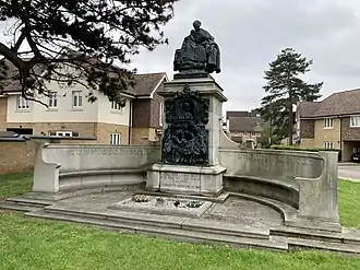 Memorial consisting of two long, curved stone seats attached to a tall stone plinth with a large bronze relief, crowned with a bronze sculpture of a woman with two children.