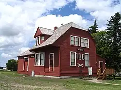 A small dirt path leads from the bottom right to the side of a red building and a five-step deck stairway at the end of which is a door. The side of the building is two stories, with two narrow windows on the bottom floor flanking the number 100, below which is a railroad crossing as a decoration and above which is a sign reading "Miami". Above these are two square windows. The building extends away to the left, with the steep roof pierced by a gable, beyond which is the roof of the single-storey freight shed. The walls consist of horizontal planks painted red, and the window frames are painted white. Behind the building are a few trees, with a cloudy sky in the background, and fronting the structure is green grass, patchy in some areas.