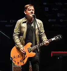 Michal Hrůza wearing black jeans and a brown leather jacket over a black t-shirt, standing onstage in front of a microphone, playing an electric guitar