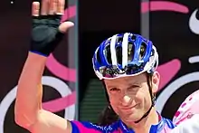 A cyclist being introduced to a crowd, waving his right hand in recognition