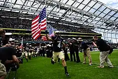 An American football player in blue and gold uniform holding a United States flag charges onto the playing field as photographers and teammates cheer.