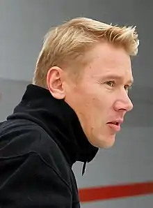 Head and shoulders of a man in his late thirties with blonde hair and grey eyes, facing to the right. He is wearing a black polo neck sweater.