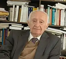 a seated portrait of an old man with a bookcase behind him