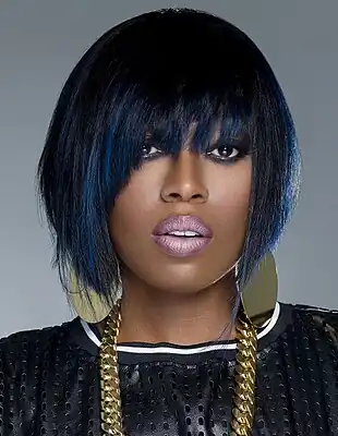 Image of rapper Missy Elliott looking to the camera with her mouth open. She is sporting short black and blue hair, heavy make-up, black shirt with white stripes, and gold earrings and necklace.