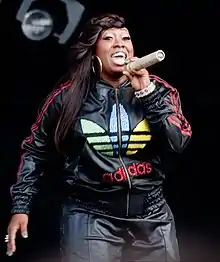 Missy Elliott posing on a grey background wearing a black shirt and gold jewelry.