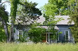 Photo of a blue one-story dogtrot-style house with enclosed breezeway sits behind overgrown trees, shrubs and grass