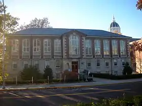 A tall brick building with a hipped roof, central pavilion and stone foundation seen from across a road with the sun just beyond the left of the image.