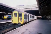 Stationary train with a yellow front and a grey, blue, white and black livery featuring the British Rail double arrow logo towards the front end.