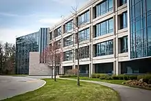 Closeup of Monster Worldwide's main headquarters, located in Weston, MA, pictured on a sunny day with blue skies. The office building is made up of large windows of blue, reflective glass framed by tan concrete. A driveway, walking path, and green landscaping are shown in front of the building.