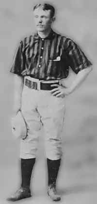 A baseball player standing, facing slightly to his right, with his right arm relaxed and holding a baseball cap, and his left arm raised so that his left hand his resting on his hip