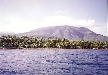 Image of a volcano that steadily rises over the horizon in the background. In the foreground is a sea, and in the middleground is a large swath of mangrove and other jungle trees.