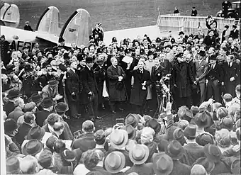 A large crowd on an airfield; British prime minister Neville Chamberlain presents an assurance from German chancellor Adolf Hitler.