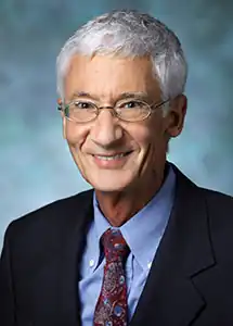 portrait photo of Roland Griffiths wearing a light blue shirt, dark blazer, and patterned tie, smiling directly at camera