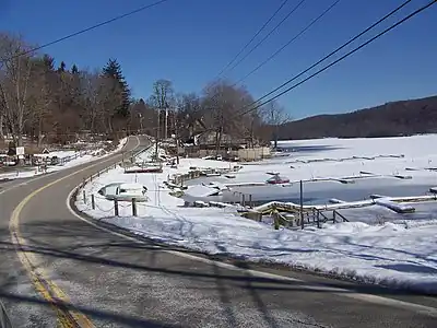 A road winding by the shore of a frozen lake and some docks in wintertime. There are telephone lines over the road, light snow on the ground, and a wooded hill on the far side of the lake at the right of the image