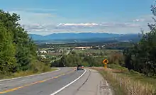 A road curving to the left with a car on it and a sign warning of a steep grade. It is descending toward a lower area with some buildings and another stretch of road. In the distance is a line of mountains.