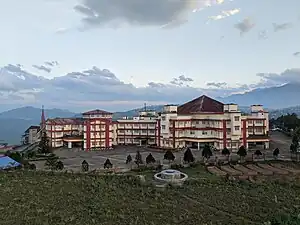 Photo of a cream-colored building with dark red highlights, with a larger section at the right capped by a brown pyramidal roof, and a narrower wing extending far off the to the left. The building is located on a hilltop with mountains in the background, and between the building and the camera lies a large, empty parking lot surrounded by a row of small decorative coniferous trees.