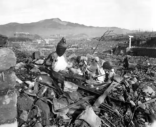 Black and white photo of a large plain covered in rubble. A badly damaged building is visible in the background.