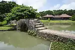 A pond crossed by a stone causeway with a bridge. There is a low wooden house beyond the pond.