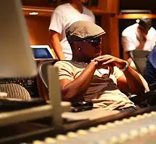 A shadowed black male wearing sunglasses and a beret sits in a recording studio, with console tools out of focus in the foreground