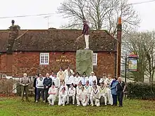 Two cricket teams plus umpires, scorers and groundsman assemble at granite monument on side of cricket pitch by the bat and ball pub for a group photo