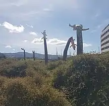 clear blue sky, foregound are plants, and long thin bending mokomoko sculptures with flapping arms