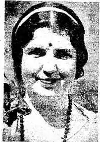 A white woman with dark hair parted center, wearing a bindi and beads, smiling.