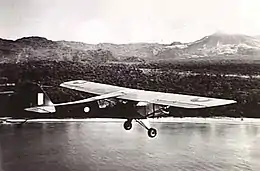 Single-engined high-wing light plane flying over water, with jungle terrain in background